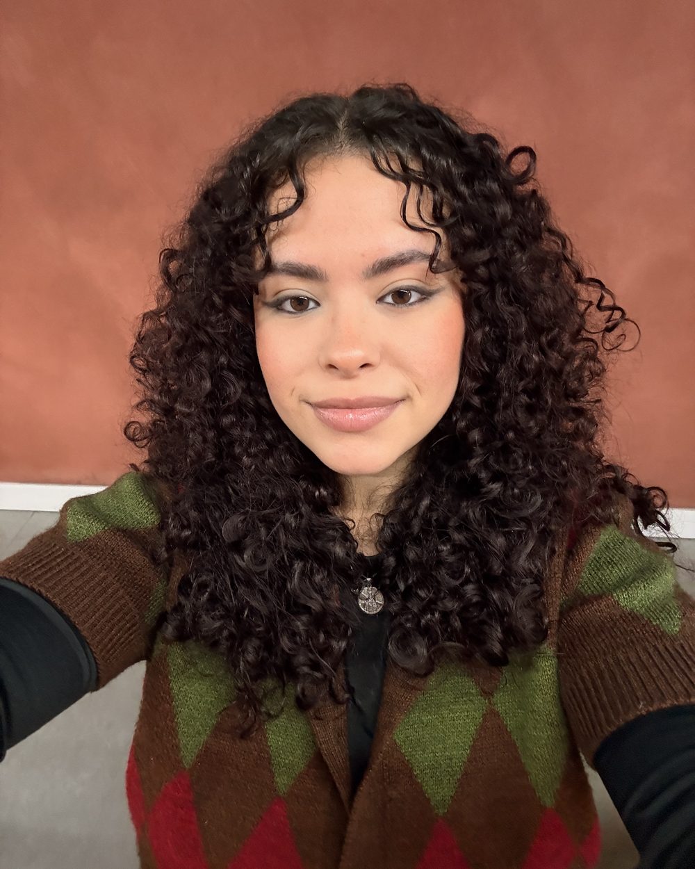 Retrato de Ingrid Lima sorrindo, com cabelo cacheado, diante de uma parede terracota